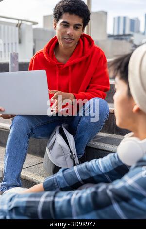 Diverse friends discussing project on laptop while sitting on rooftop steps near gray backpack Stock Photo