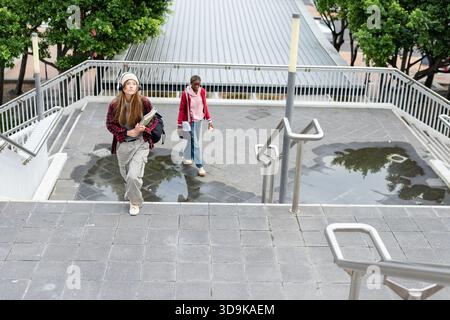 Diverse female friends walking up campus stairs holding notebooks and carrying backpack coffee cup Stock Photo