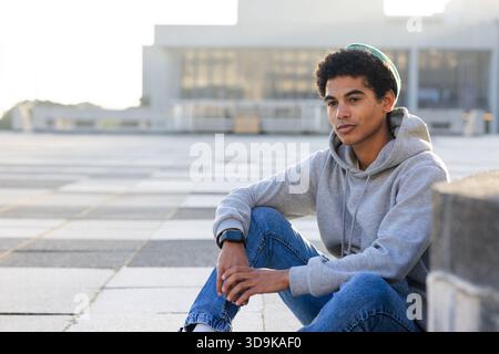 Hispanic male student sitting on stone ledge at urban plaza tiles, checking smartwatch, copy space Stock Photo