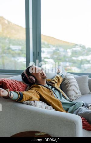 Mid adult man relaxing on sofa with brick red pillow listening through wireless over ear headphones Stock Photo