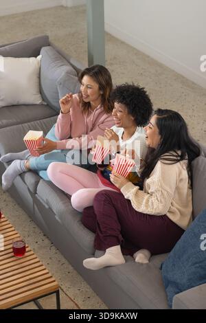 Women sitting on gray sectional sofa at home wearing loungewear and socks holding popcorn tubs Stock Photo