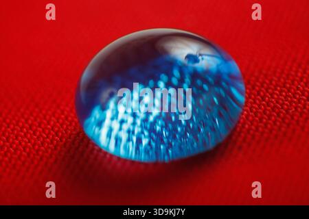 A dark blue liquid droplet rests perfectly spherical on a bright red, tightly woven, water-repellent fabric, highlighting surface hydrophobicity. Stock Photo