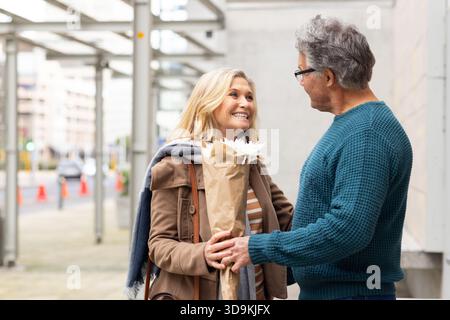 Senior couple standing on sidewalk facing each other holding bouquet of white daisies and handbag Stock Photo