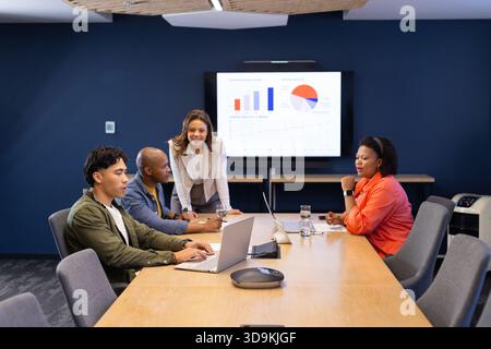 Diverse coworkers sitting around table in conference room using laptops and viewing chart display Stock Photo