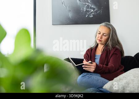 Senior woman writing with pen in black notebook on sofa by window, blurred houseplant, copy space Stock Photo