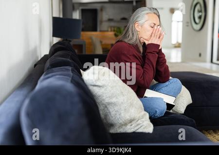 Senior woman sitting on dark-blue sofa in open-plan home pressing hands to face holding closed book Stock Photo