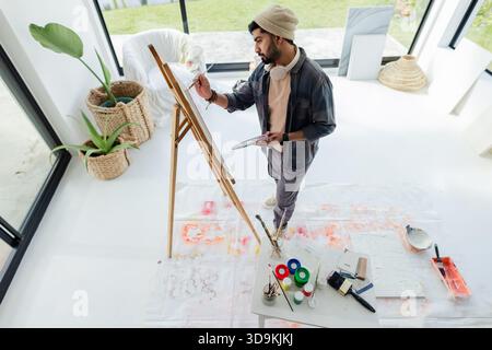 Indian man painting on canvas at easel in bright home studio with paint jars and palette Stock Photo