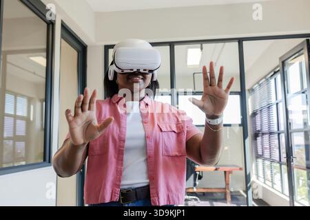 Adult woman wearing white VR headset raising hands and engaging with virtual display inside office Stock Photo