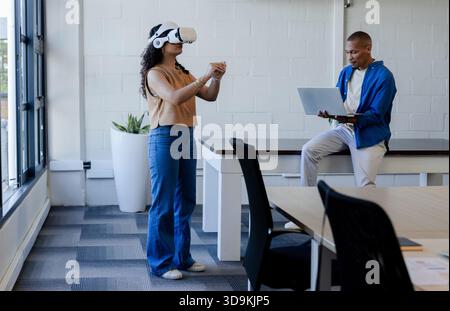 African American casual team testing VR by windows, woman gesturing as man using laptop Stock Photo