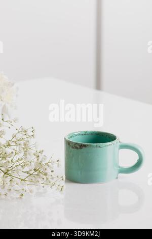 A beautiful handmade turquoise ceramic cup beside a bunch of white flowers on a clean white counter. The image has negative space to write some text. Stock Photo