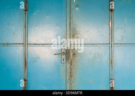 Close-up of a weathered and rusty blue metal double door with strong geometric panel divisions. The aged surface features peeled paint, rust stains Stock Photo