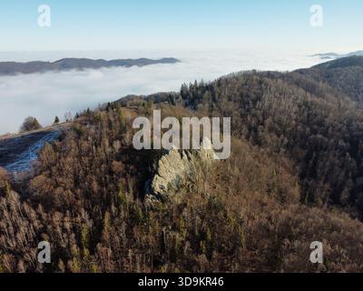 Aerial view of rugged rock formations amidst dense, leafless forest. Scene bathed in crisp daylight, with rolling hills and sea of clouds in background. Carpathian mountains, Ukraine. Stock Photo