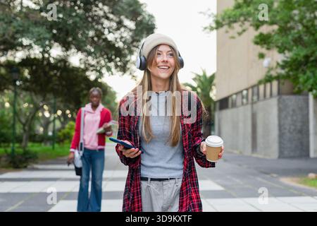 Diverse female students walking on campus smiling with smartphone and coffee cup wearing headphones Stock Photo
