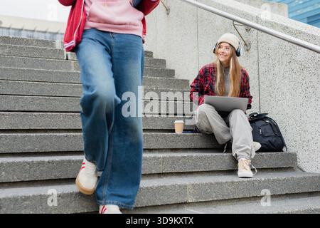 Female students studying on concrete steps outside glass building using laptop with coffee cup Stock Photo