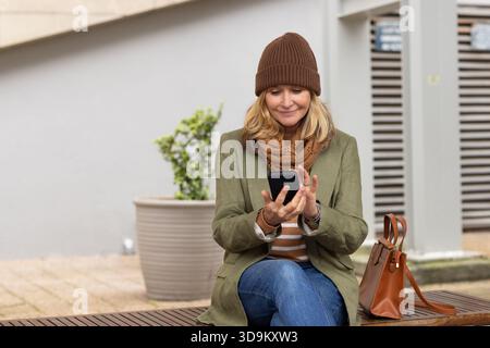 Woman sitting on bench in urban plaza, using smartphone with brown handbag beside Stock Photo