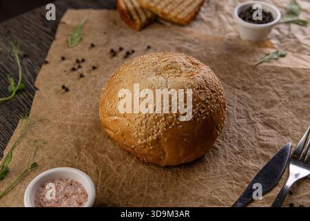 Round sesame seed bun placing on crinkled craft paper with seasonings and cutlery Stock Photo