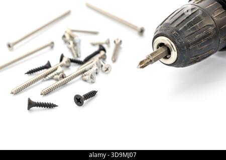 A close-up view of a black cordless drill positioned beside various screws on a clean white background. The drill shows a metallic bit, ready for use, Stock Photo