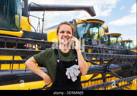 Female agricultural machinery mechanic talking on mobile phone against combine harvesters. Stock Photo