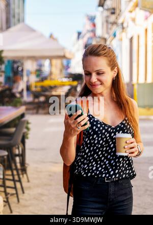 Smiling urban woman with paper coffee cup using her smartphone on the go. Stock Photo