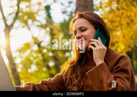 Attractive female talking mobile phone while working on laptop outdoors. Stock Photo