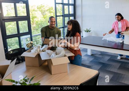 Diverse coworkers unpacking framed picture and bubble wrap at wooden tables in renovated office Stock Photo