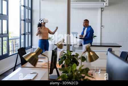 African American coworkers showing VR headset demo while using laptop in casual office with windows Stock Photo