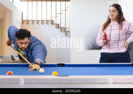 Couple leaning over blue pool table aiming cue stick at home rec room, holding bottled drink Stock Photo