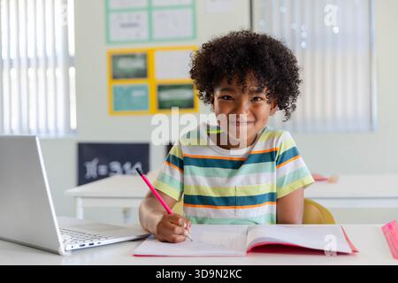 African male youth student sitting at class desk writing in notebook with pink pencil, striped tee Stock Photo