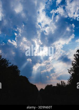 Dramatic sky with glowing sunbeams breaking through dense clouds above a silhouetted landscape and railway lines, creating a powerful atmospheric scen Stock Photo