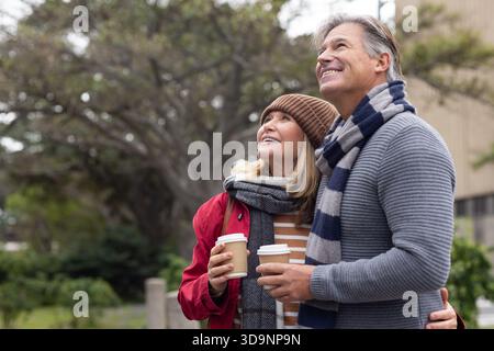 Couple standing in park wearing scarves and beanie holding takeaway coffee cups looking upward Stock Photo
