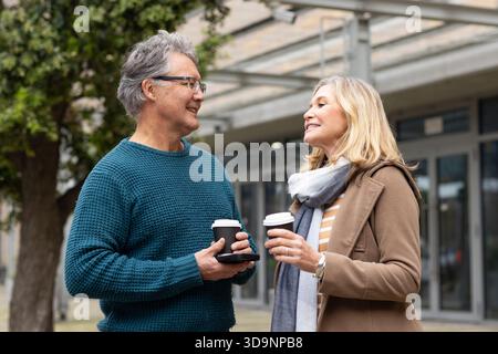 Senior couple holding coffee cups and using smartphone standing on plaza outside modern building Stock Photo