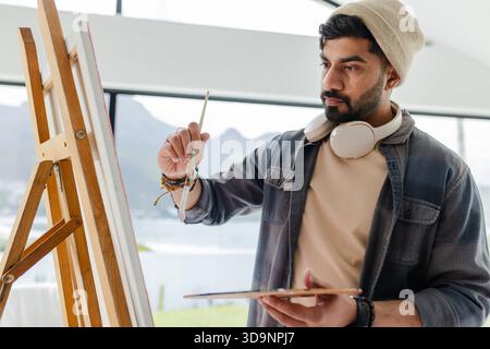 Indian male artist standing at easel in bright studio painting canvas with brush palette Stock Photo