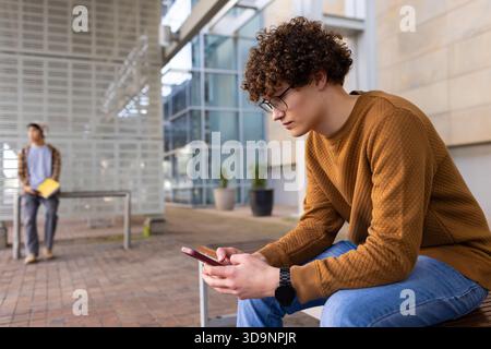 Male students sitting on campus bench using red smartphone with yellow notebook near potted plants Stock Photo