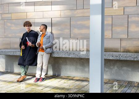 Diverse male friends standing and chatting against wood wall holding diploma portfolio, copy space Stock Photo