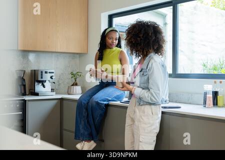 Diverse female friends chatting and holding mugs on home kitchen countertop near espresso machine Stock Photo