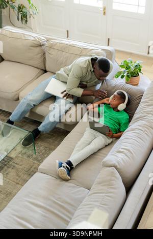 African American man and boy sitting on sectional sofa at home using laptop tablet white headphones Stock Photo