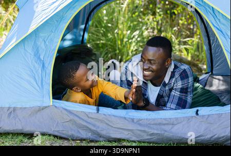 African American father and son high-fiving in blue-grey tent with green sleeping bag and backpack Stock Photo