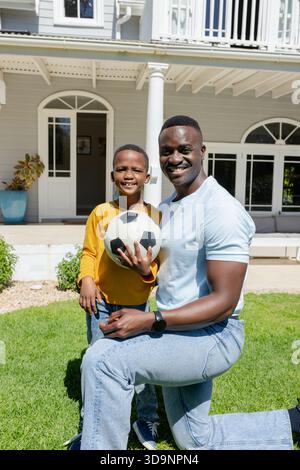 African American father and son kneeling, holding soccer ball on lawn in front of house Stock Photo