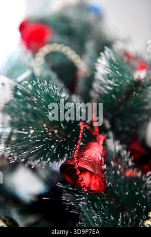 Vertical close-up of a frosted artificial Christmas tree branch featuring a bright red metallic bell ornament and a delicate gold bead garland. Stock Photo