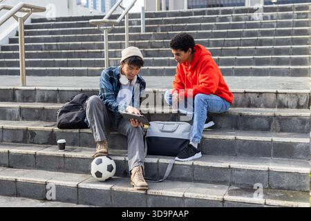 Diverse male students sitting on campus steps reviewing tablet with soccer ball and coffee cup Stock Photo