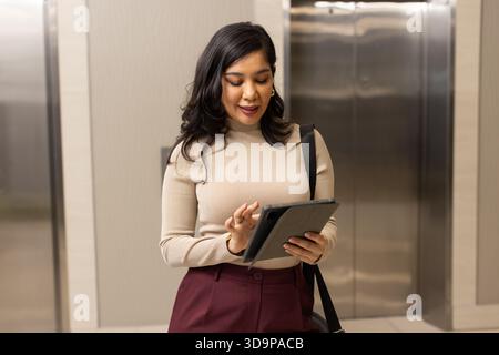 Hispanic or Latino woman tapping tablet, carrying bag by elevator call button panel in office lobby Stock Photo