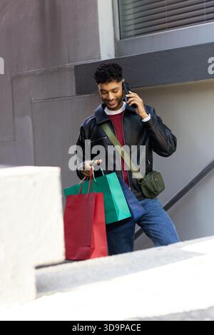 Man holding three colorful shopping bags and speaking on smartphone by handrail on outdoor stairs Stock Photo