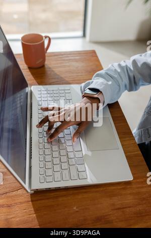 Hand with pink smartwatch is typing on white laptop at wooden desk, sunlight casting shadows Stock Photo