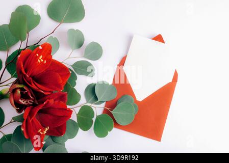 Red amaryllis flowers and green eucalyptus leaves beside an orange envelope with blank card. Top view, flat lay, mockup. Stock Photo
