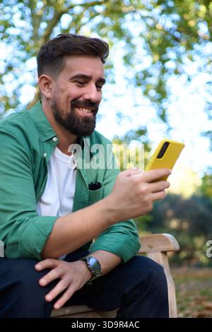 Smiling man on park bench using yellow smartphone in autumn Stock Photo