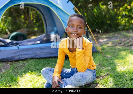 African American boy sitting on yard grass in yellow shirt facing blue-gray tent with sleep pad Stock Photo