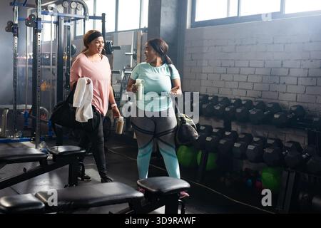 Diverse mid adult women in workout clothes holding water bottles by dumbbell rack in gym Stock Photo