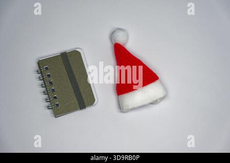 A small spiral notebook lies beside a red Santa hat on a clean white background, creating a simple festive scene ideal for holiday planning, Christmas Stock Photo