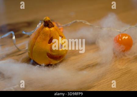 Small ceramic pumpkin Halloween decoration on wooden table with cotton and glowing pumpkin lights creating festive mood Stock Photo