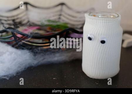 A jar wrapped in gauze with two plastic eyes placed on a black marble table, with cotton decoration and a perforated black glass dish in the backgroun Stock Photo